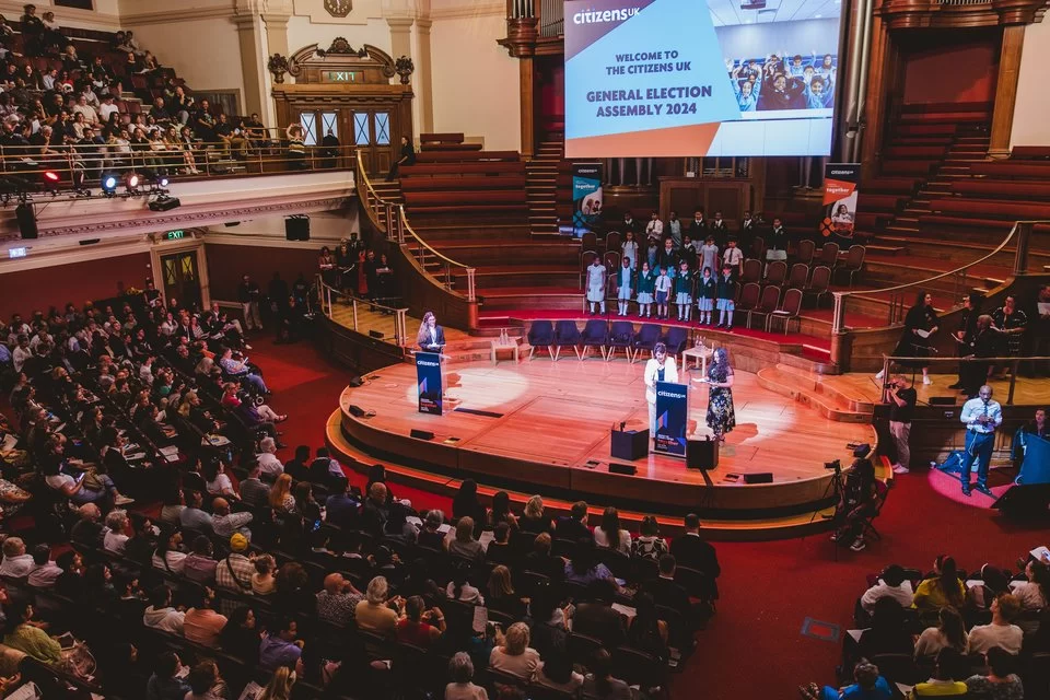A view from above the Citizens Assembly. An audience sitting in rows looking at figures on stage. A presentation screen with the words: "Welcome to the Citizens UK General Election Assembly 2024"